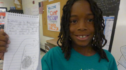 A youth smiling while holding up a notepad in a classroom