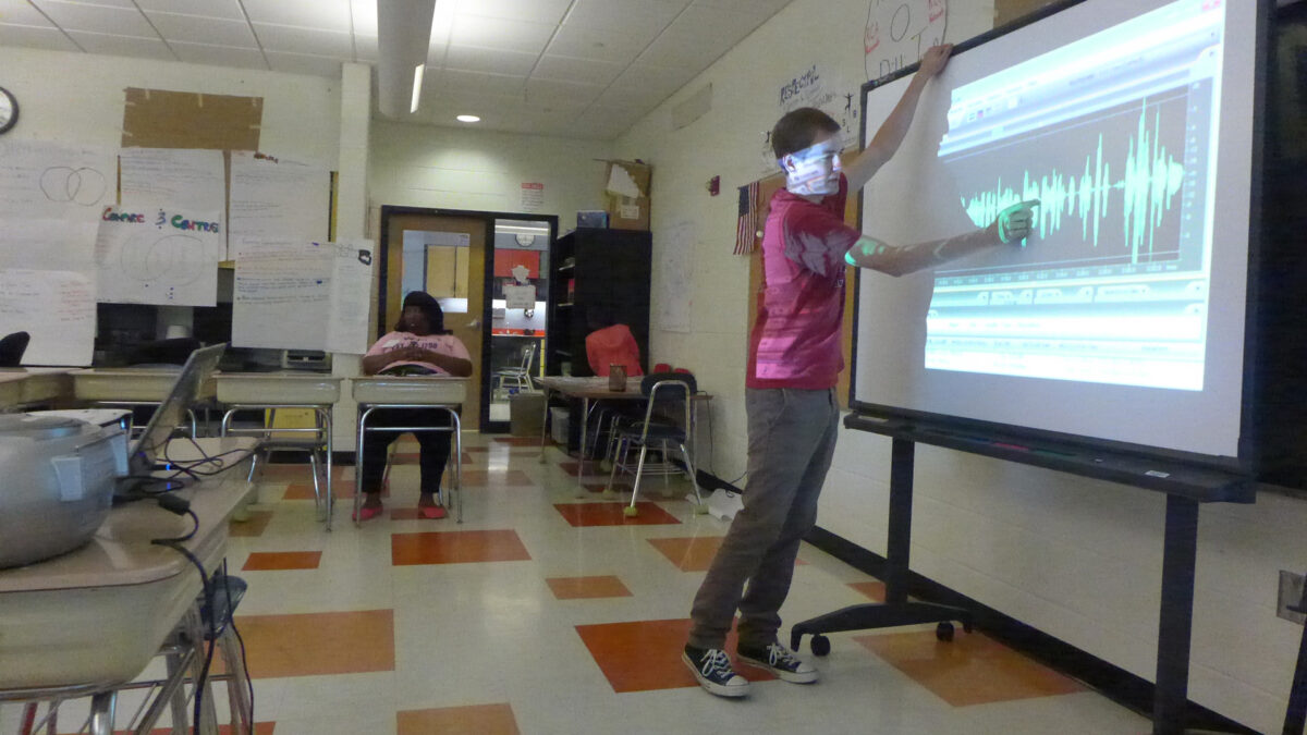An adult gesturing at audio editing software on a projected screen in a classroom
