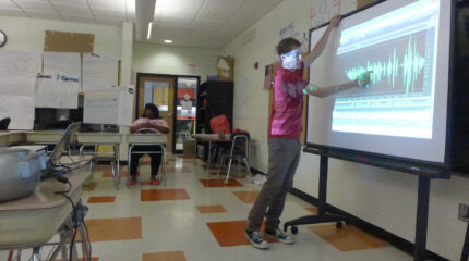 An adult gesturing at audio editing software on a projected screen in a classroom