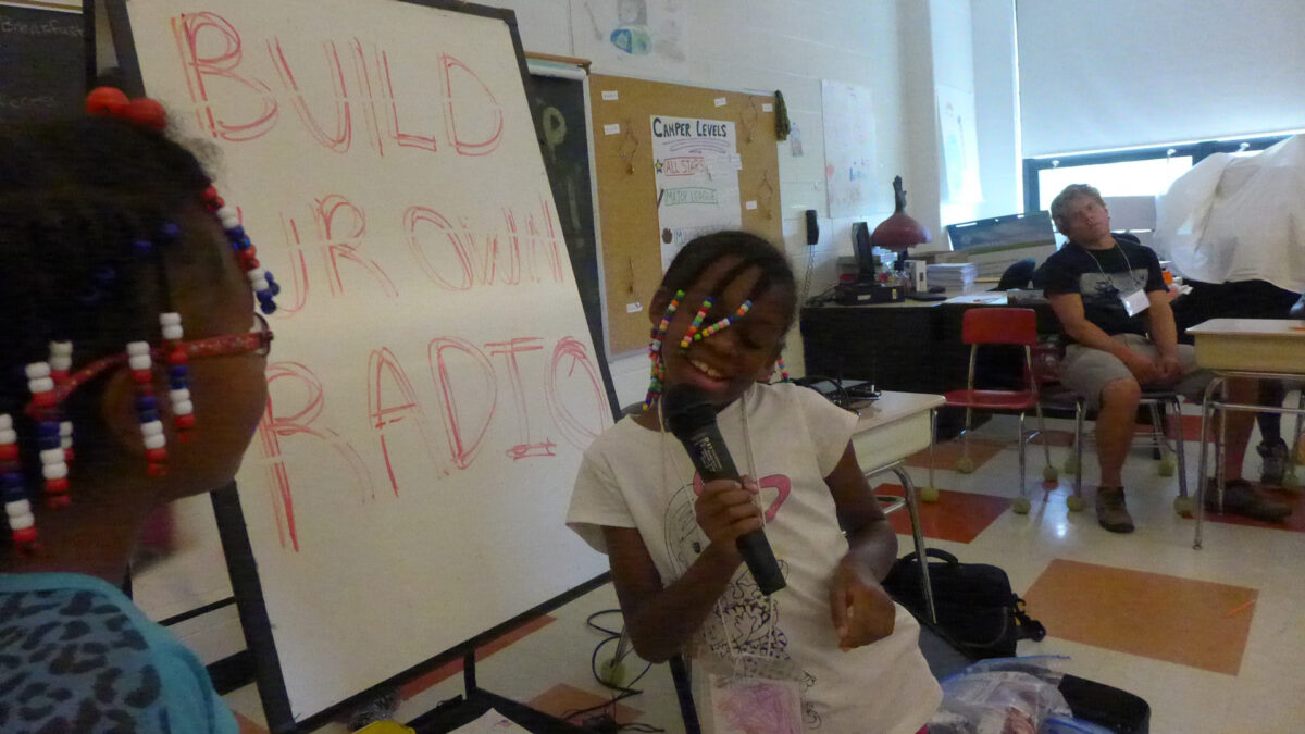 A youth holding and speaking into a microphone standing in front of a white board as a youth and an adult look on in a classroom