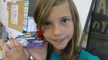A youth holding an electrical device up to their ear and smiling in a classroom
