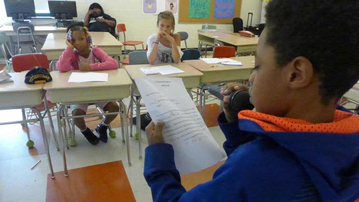 A youth looking at a piece of paper and speaking into a held microphone as three youth listen while sitting at classroom desks