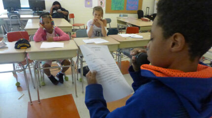 A youth looking at a piece of paper and speaking into a held microphone as three youth listen while sitting at classroom desks