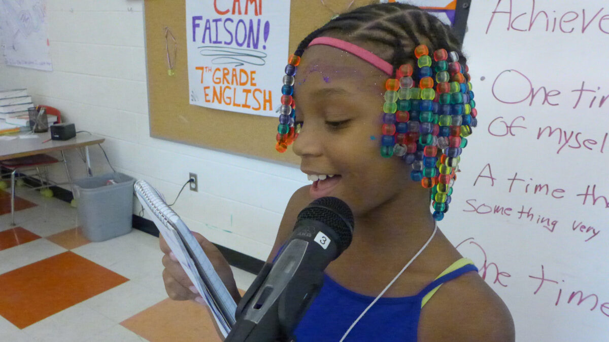A youth reading a notepad and speaking into a microphone standing in front of a whiteboard in a classroom