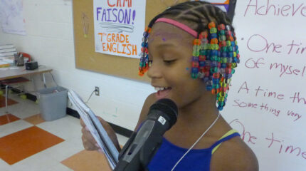 A youth reading a notepad and speaking into a microphone standing in front of a whiteboard in a classroom