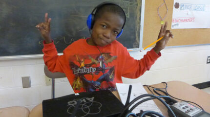 A youth wearing headphones connected to a portable recorder sitting at a classroom desk and flashing two peace signs with their hands in front of a blackboard