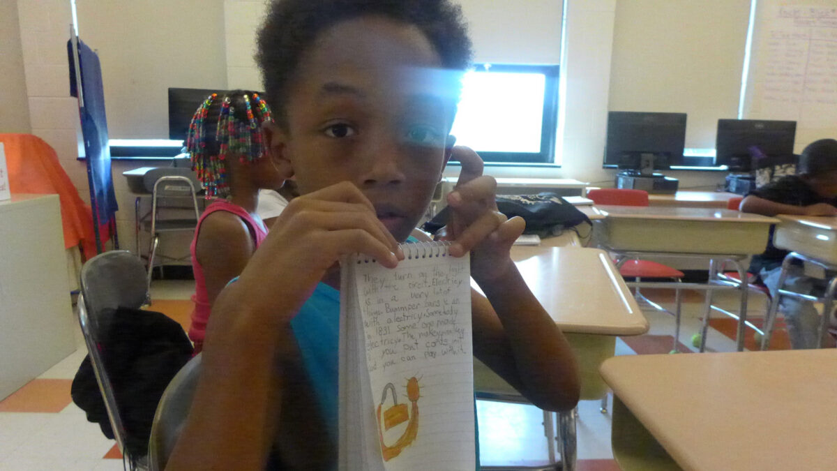 A youth holding a notepad up to the camera and another youth sitting at a classroom desk in the background