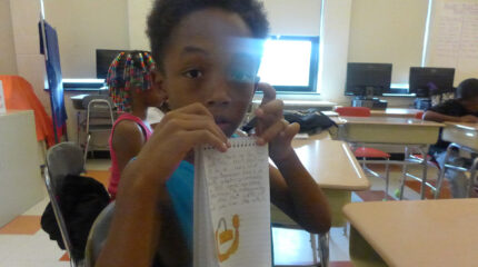 A youth holding a notepad up to the camera and another youth sitting at a classroom desk in the background
