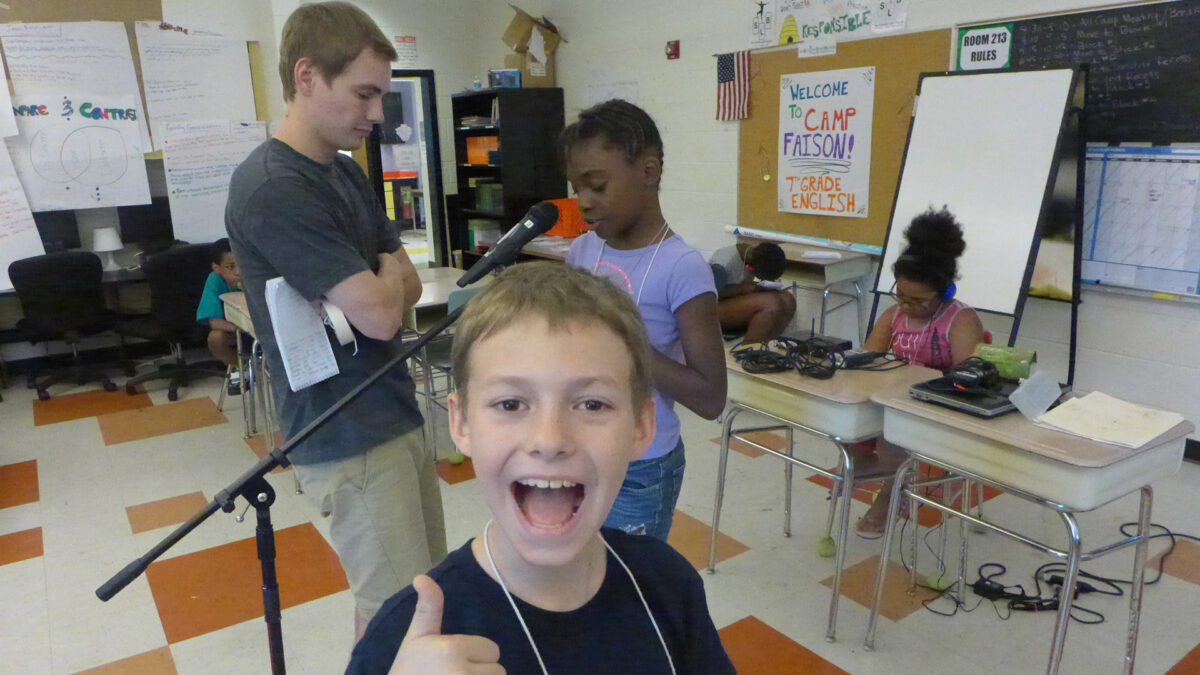 A youth giving a thumbs up standing in front of an adult looking at a youth speaking into a microphone and a youth wearing headphones sitting in the background at a classroom desk looking at a portable recorder