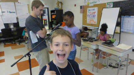 A youth giving a thumbs up standing in front of an adult looking at a youth speaking into a microphone and a youth wearing headphones sitting in the background at a classroom desk looking at a portable recorder