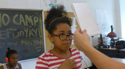 A youth looking at a notepad held in front of their face and a youth wearing headphones sitting in front of a chalkboard