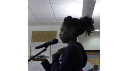 A youth holding a cell phone and speaking into a microphone in a classroom
