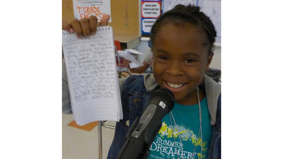 A youth smiling while standing behind a microphone and holding up a notepad in a classroom