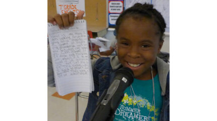 A youth smiling while standing behind a microphone and holding up a notepad in a classroom