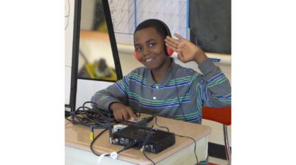 A youth wearing headphones and giving a salute while sitting behind a portable microphone on a classroom desk