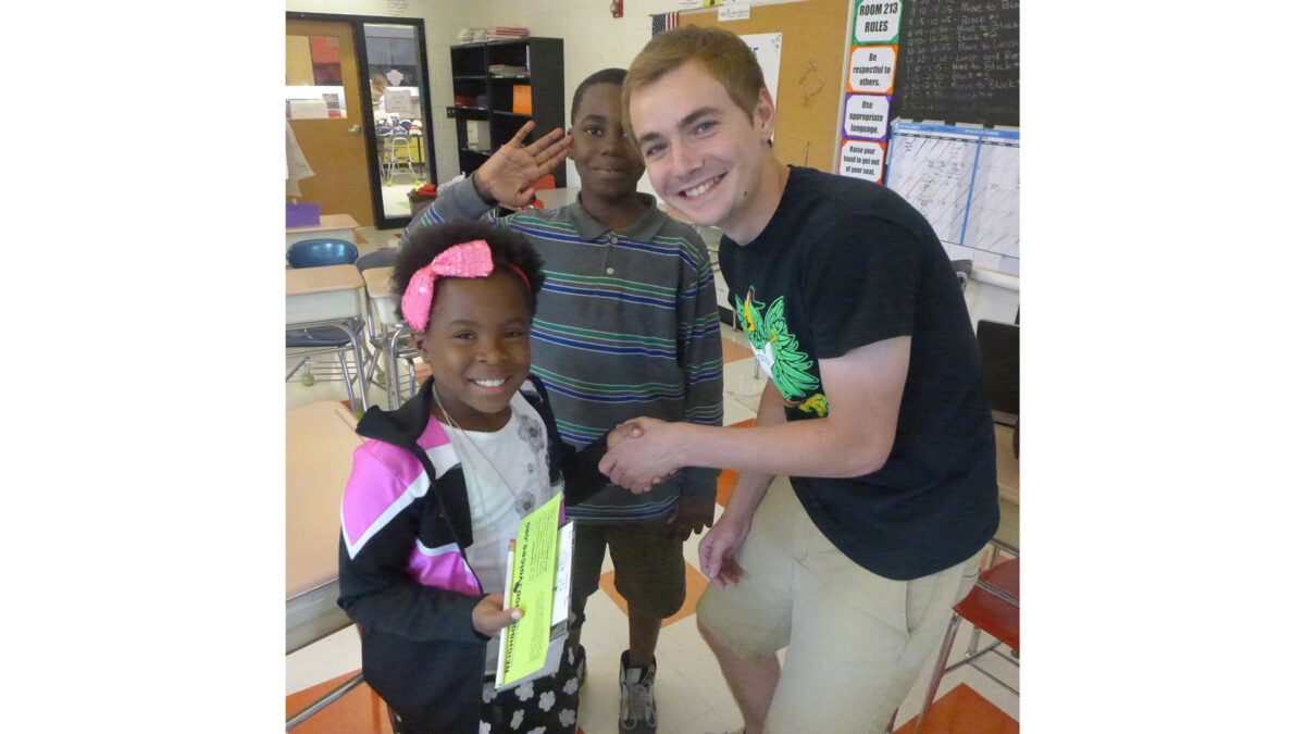 An adult and a youth shaking hands while another youth salutes behind them in a classroom