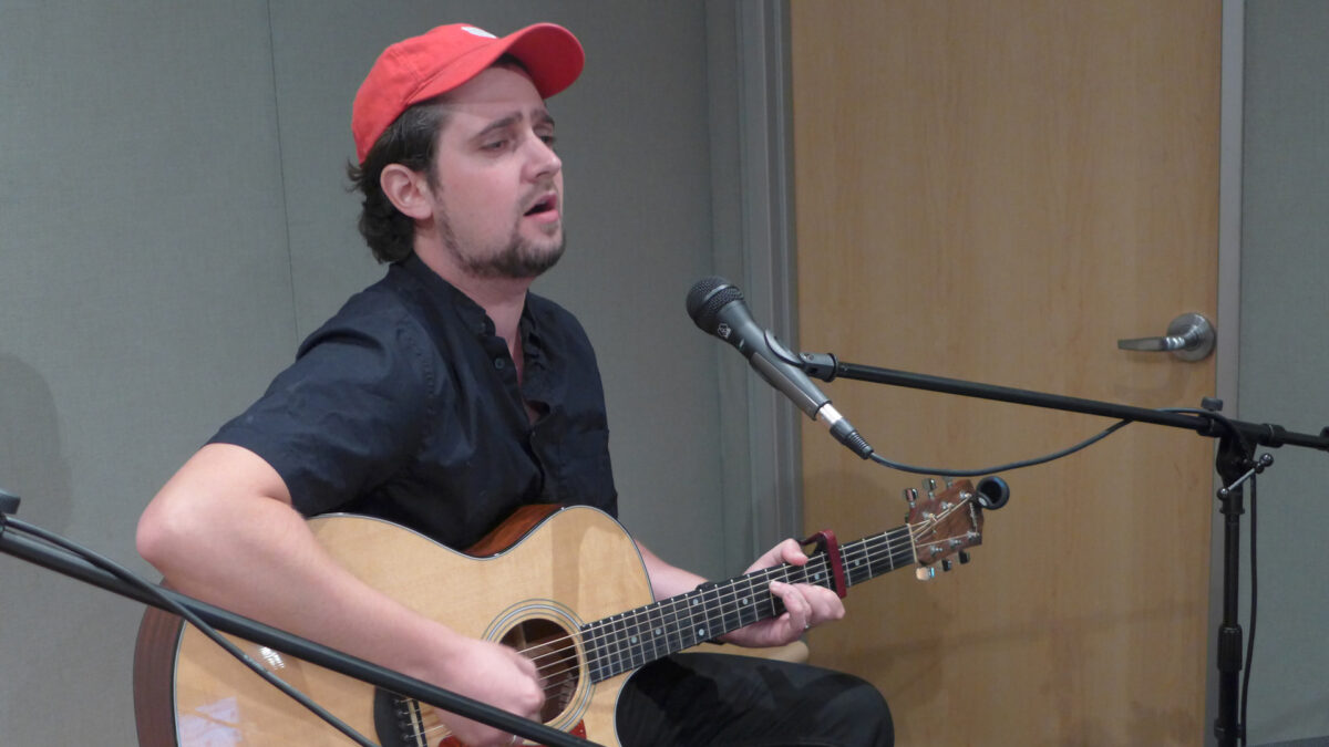 An adult playing an acoustic guitar and singing into a microphone in a radio studio