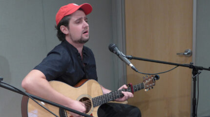 An adult playing an acoustic guitar and singing into a microphone in a radio studio