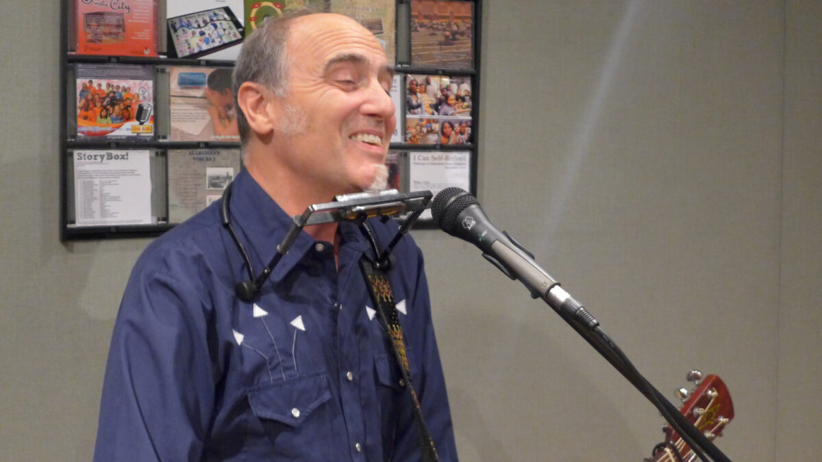 An adult with a harmonica around their neck smiling while holding an acoustic guitar and singing into a microphone in a radio studio