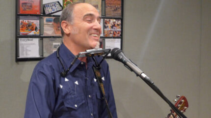 An adult with a harmonica around their neck smiling while holding an acoustic guitar and singing into a microphone in a radio studio