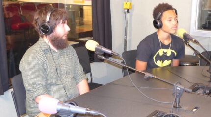 An adult and a teen wearing headphones speaking into microphones in a radio studio