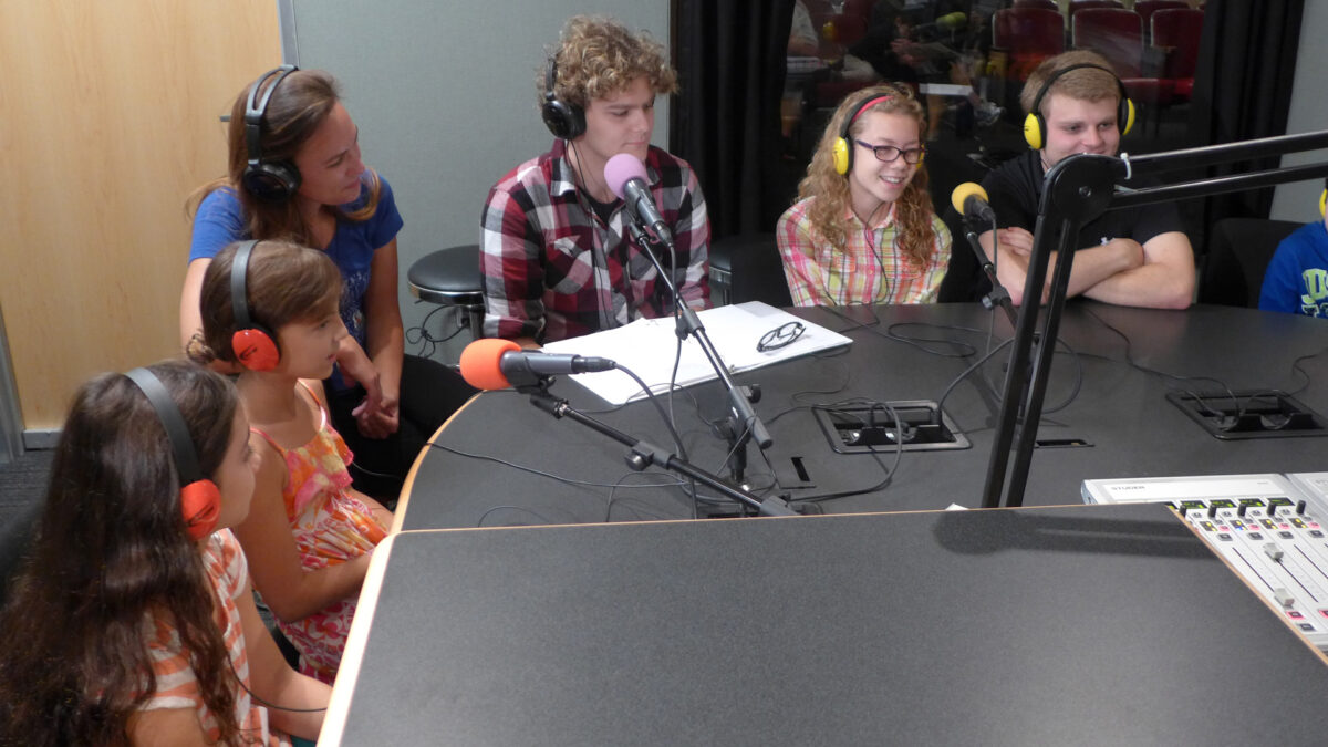 Seven youth wearing headphones and speaking into microphones in a radio studio