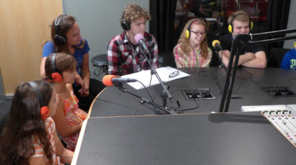 Seven youth wearing headphones and speaking into microphones in a radio studio