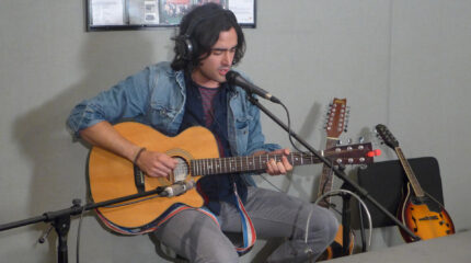 An adult wearing headphones playing an acoustic guitar and singing into a microphone in a radio studio