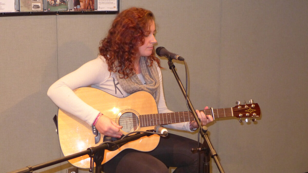 A teen playing acoustic guitar sitting behind a microphone in a radio studio