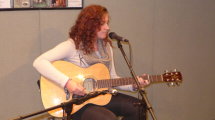 A teen playing acoustic guitar sitting behind a microphone in a radio studio