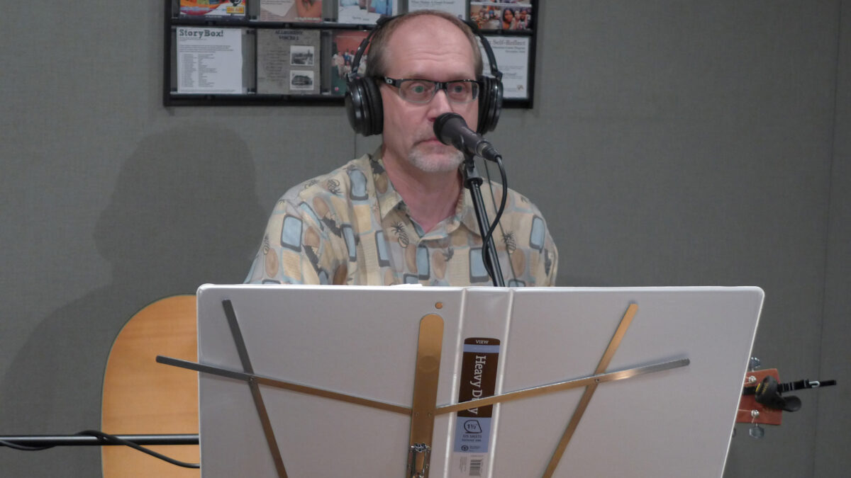 An adult wearing headphones sitting behind a microphone and a music stand with a white binder on it in a radio studio