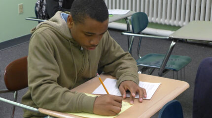 A teen writing on a piece of paper on a classroom desk