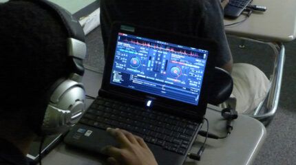 An individual wearing headphones looking audio mixing software on a laptop computer sitting at a classroom desk