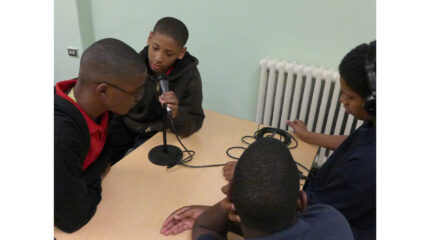 Four teens sitting around a wooden table, one speaking into a microphone, one wearing headphones