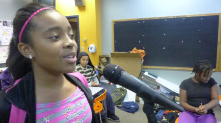A youth speaking into a microphone and two youth sitting in a classroom