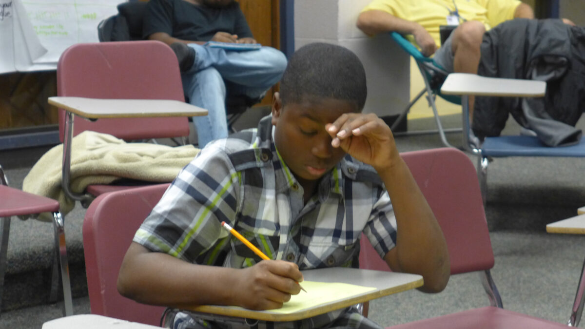 A youth looking down and writing on a piece of paper on a classroom desk in a classroom