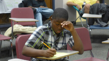 A youth looking down and writing on a piece of paper on a classroom desk in a classroom