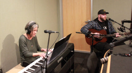 An adult wearing headphones playing a piano keyboard and singing into a microphone and an adult playing acoustic guitar in a radio studio