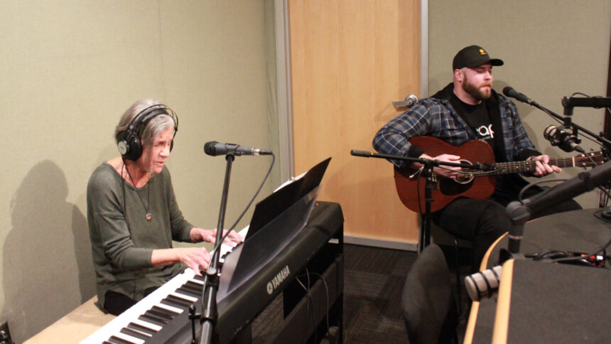 An adult wearing headphones playing a piano keyboard and singing into a microphone and an adult playing acoustic guitar in a radio studio
