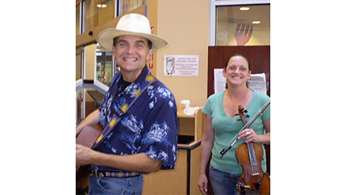 An adult holding an acoustic guitar and an adult holding a violin, both smiling at the camera