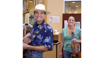An adult holding an acoustic guitar and an adult holding a violin, both smiling at the camera