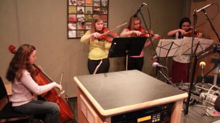 A teen playing cello, two teens playing violin, and a teen playing viola, all looking at sheet music on music stands in a radio studio