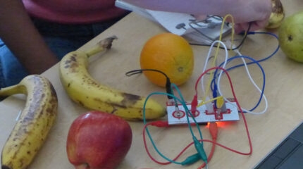 Two bananas, an apple, an orange, and a white rectangle with wires coming out, all on a classroom desk