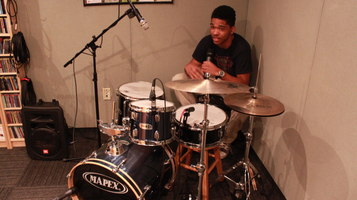 A teen holding and speaking into a microphone while sitting at a drum set in a radio studio