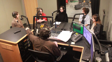 Five youth wearing headphones speaking into microphones being interviewed by an adult wearing headphones sitting behind a microphone and a mixer in a radio studio