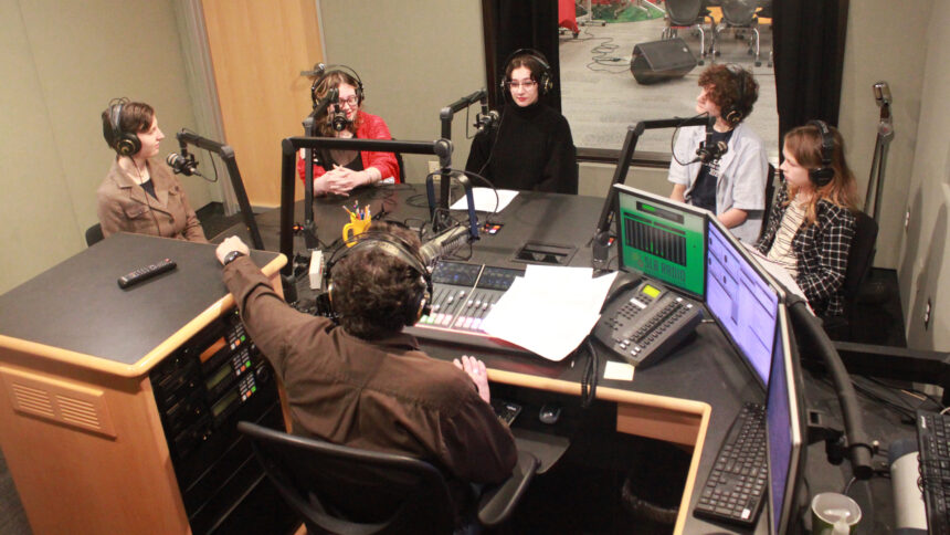 Five youth wearing headphones speaking into microphones being interviewed by an adult wearing headphones sitting behind a microphone and a mixer in a radio studio