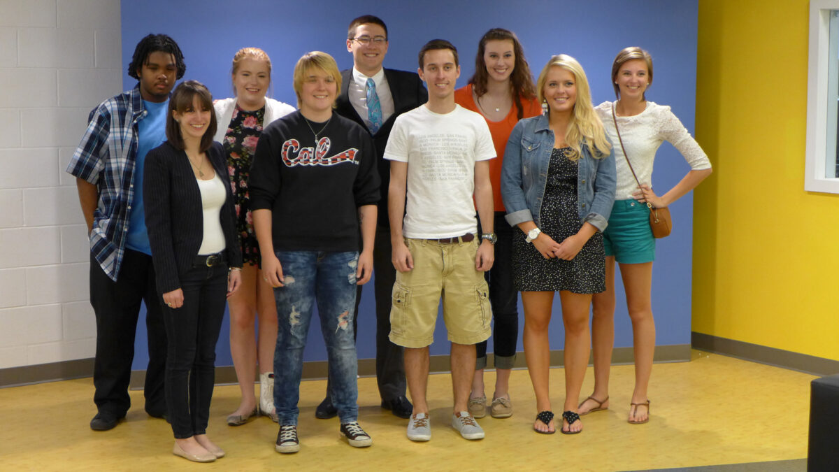 Nine adults smiling and standing in front of a blue wall