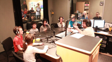 Six youth wearing headphones sitting behind microphones being interviewed by an adult wearing headphones speaking into a microphone and sitting behind a mixer in a radio studio