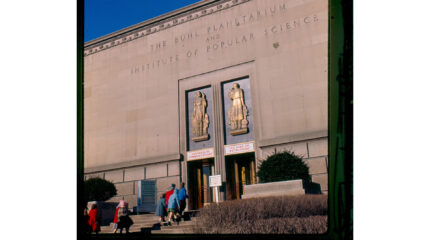Individuals walking into a large beige building with two gold statues above two doors, and the text "The Buhl Planetarium and Institute of Popular Science" engraved on the building