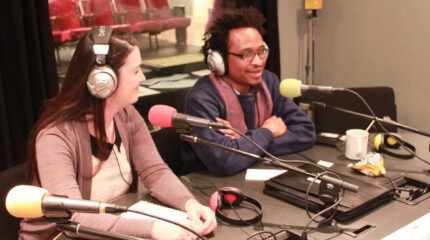 Two adults wearing headphones speaking into microphones in a radio studio